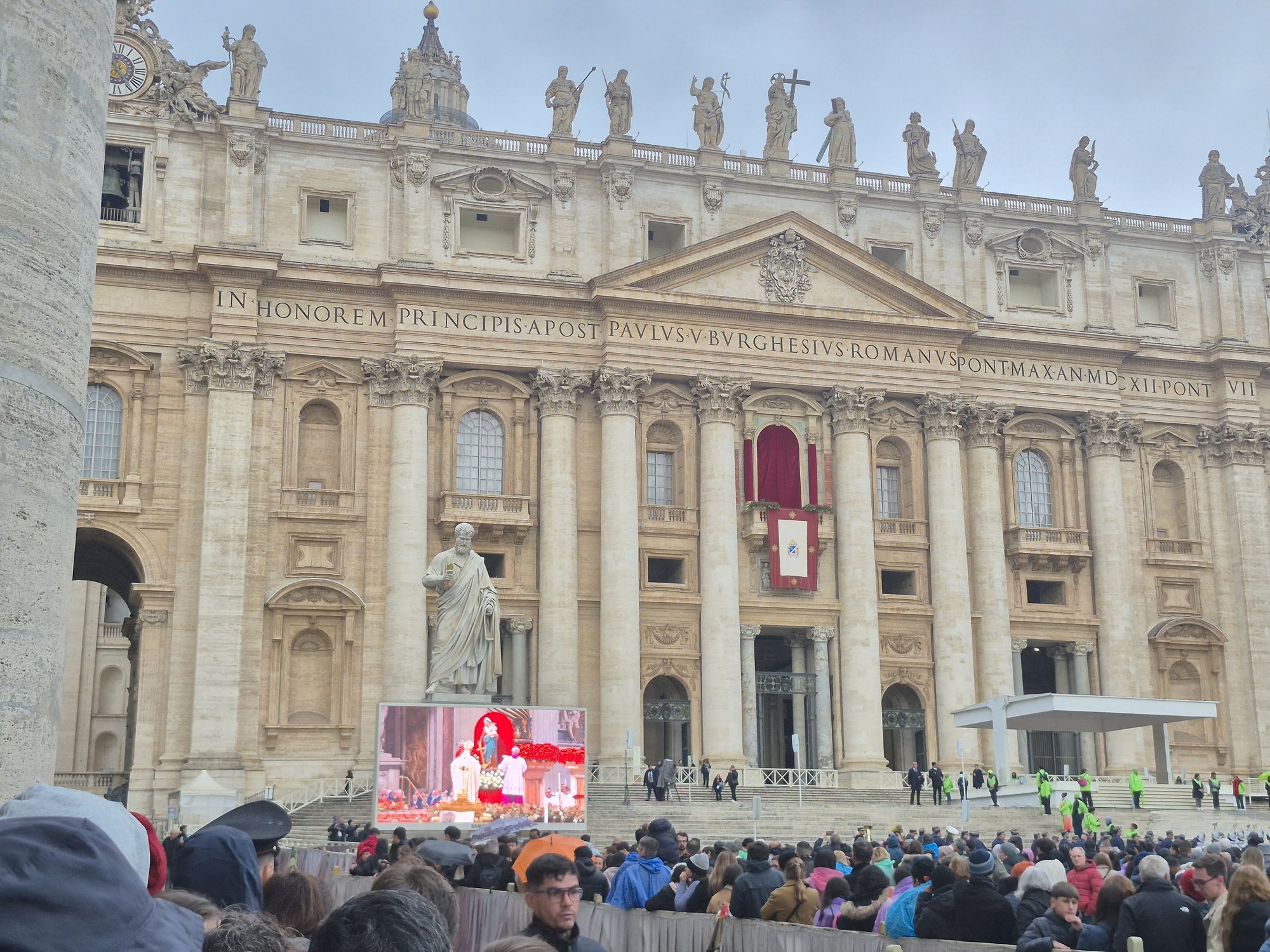 The façade of St. Peter's Basilica on Christmas morning, the papal balcony hung with red, a crowd under umbrellas in the square and the big screens showing the Mass