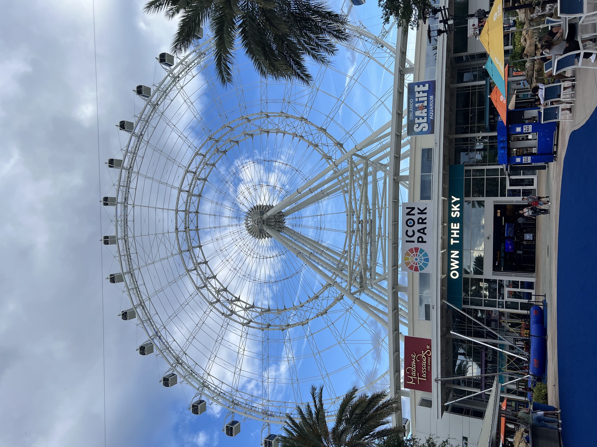The ICON Park observation wheel in Orlando against a blue sky with scattered clouds, the entrance plaza below