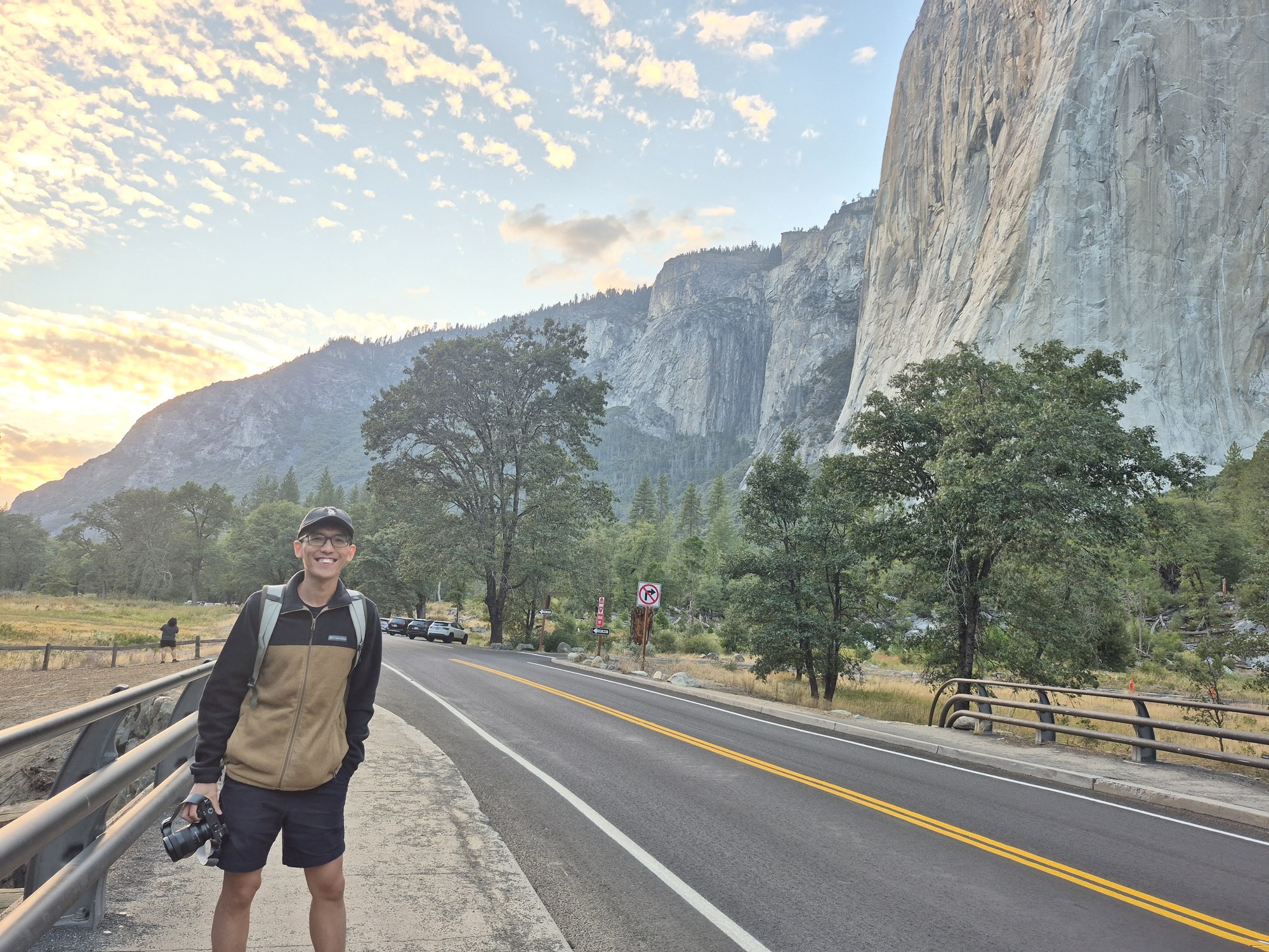 A hiker with a backpack and cap standing at the roadside guardrail in El Capitan Meadow, the pale granite face of El Capitan towering directly above through the pines